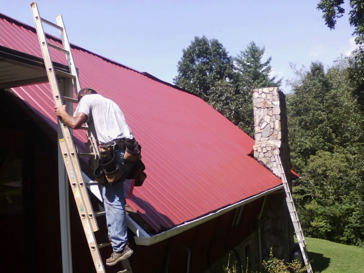 Roofer on a ladder beside a red metal roof and stone chimney — Soffit & Fascia Repair by Roofing Brothers in Mount Saint Joseph